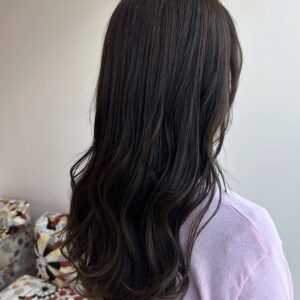 Back view of a person with long, dark brown hair in a light purple sweater, standing in a room with patterned cushions in the background.