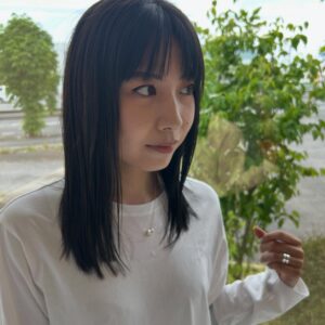 Portrait of a woman with straight black hair and bangs, wearing a white top, looking to her left beside a window with greenery outside.
